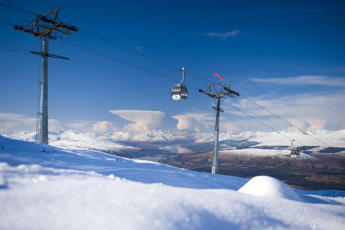 A ski gondola is seen suspended above a landscape of snow, with mountains in the background and a clear blue sky. The tracks and pylons are positioned prominently, while gentle snowdrifts create a serene foreground.