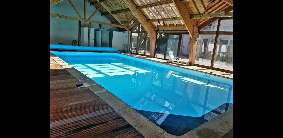 A covered pool area features a crystalline blue pool with smooth tile edges. Natural light floods through large windows, accentuating the wooden beams and rustic decor. Lounge chairs are arranged beside the pool, creating a relaxing environment for guests.