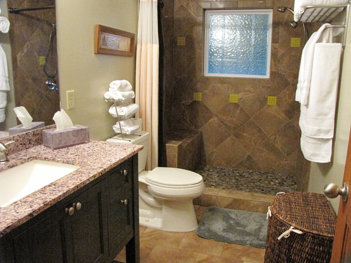 The bathroom features a spacious walk-in shower with textured glass blocks, accented by earth-tone tiles. A dark vanity with a granite countertop complements the setup, while neatly arranged towels and toiletries provide convenience. A woven basket adds a natural touch to the decor.