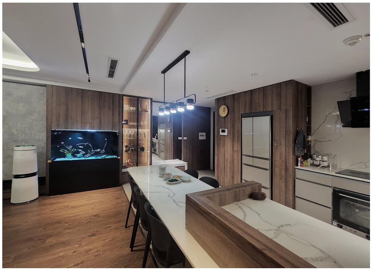 A modern kitchen area features a large marble countertop with seating. An aquarium adds an inviting touch. The space is illuminated by stylish pendant lighting. Sleek cabinetry and kitchen appliances are visible, creating a functional and welcoming environment.