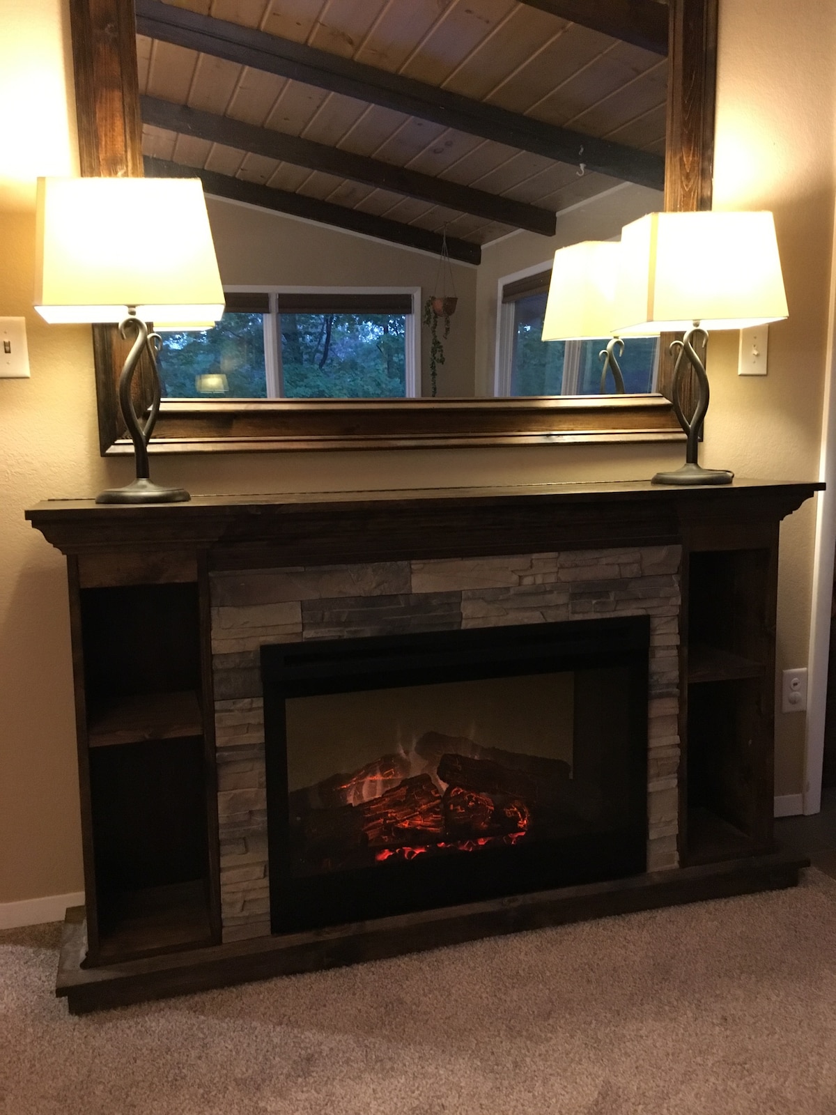 A rustic fireplace is centered beneath a large mirror, featuring a realistic flame effect. Two lamps with soft light stand on either side of the mantel, enhancing the inviting atmosphere. Shelving on the sides provides space for decorative items or personal belongings.
