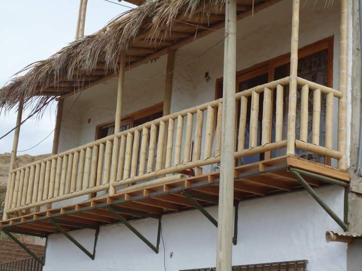 A spacious balcony with a bamboo railing extends from the second floor, showcasing wooden beams and a thatched roof. Large windows are present, allowing natural light to illuminate the area. The exterior features a rustic white wall and a sturdy support structure.