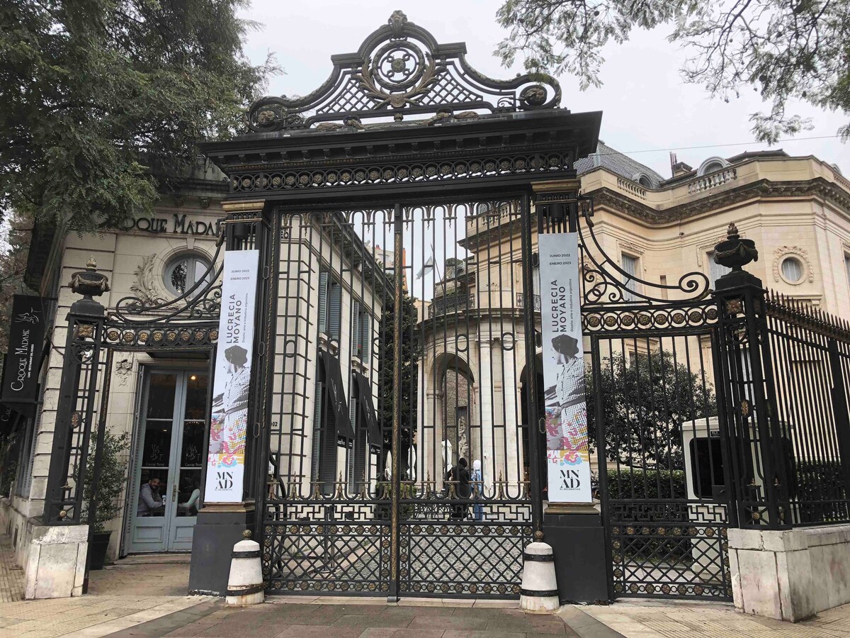 An ornate wrought iron gate, with intricate detailing, leads into a refined building. The gate features decorative elements and is flanked by vibrant banners. The surrounding area includes greenery and highlights the architectural style of the structure beyond the gate.