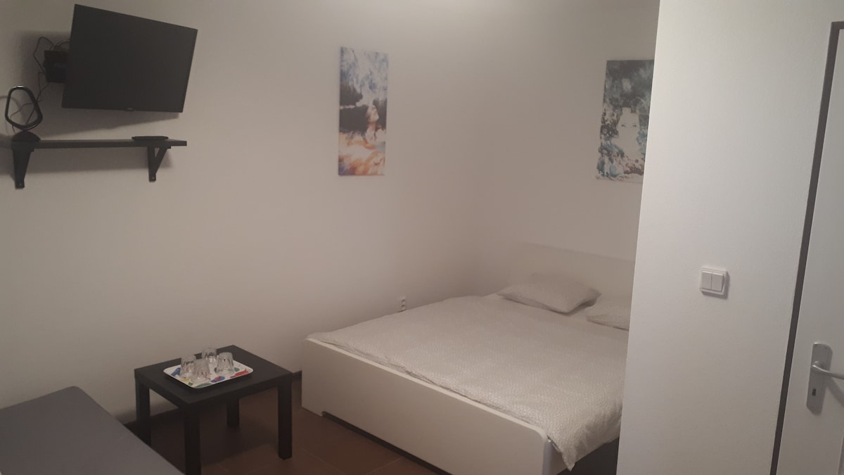 A simple and functional room features a spacious bed with light bedding. A small table is set with glassware. A flat-screen TV is mounted on the wall, and small wall art pieces enhance the ambiance, offering a calm atmosphere.