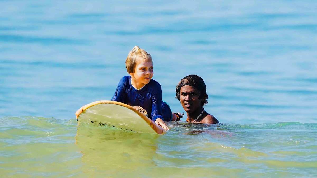 A child is seen holding a surfboard in shallow water, with an adult nearby offering support. The ocean is calm, with gentle waves and a serene blue background that emphasizes the peaceful beach setting.