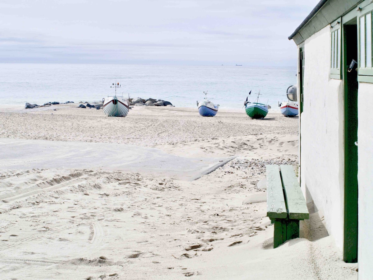 A sandy beach landscape is visible, featuring several fishing boats in varying colors along the shore. A simple wooden bench is positioned adjacent to a modest building, with the peaceful ocean waves creating a serene backdrop in the distance.