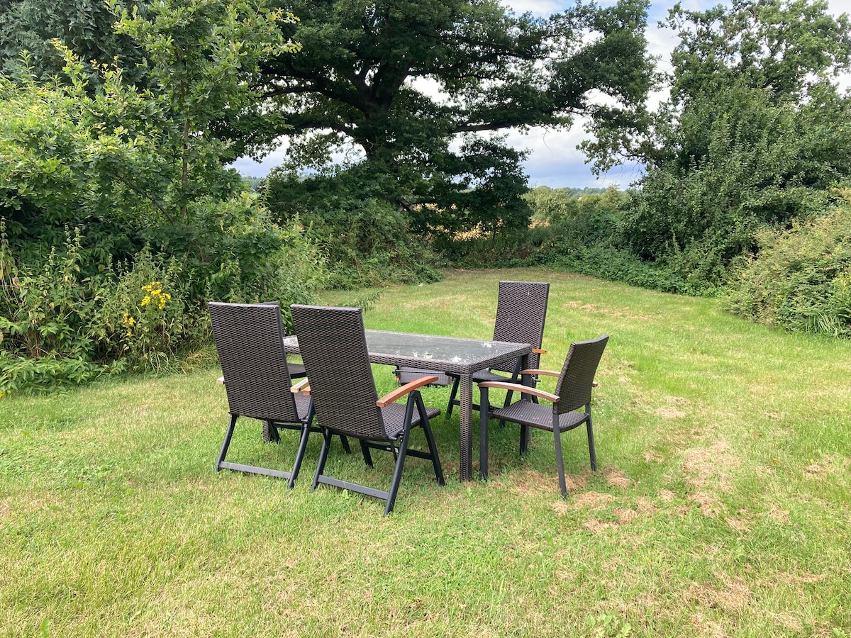 An outdoor seating area is presented with a rectangular table accompanied by four chairs. The setting is surrounded by lush greenery and tall grass, offering a peaceful environment. The sky is partly cloudy, enhancing the natural atmosphere.