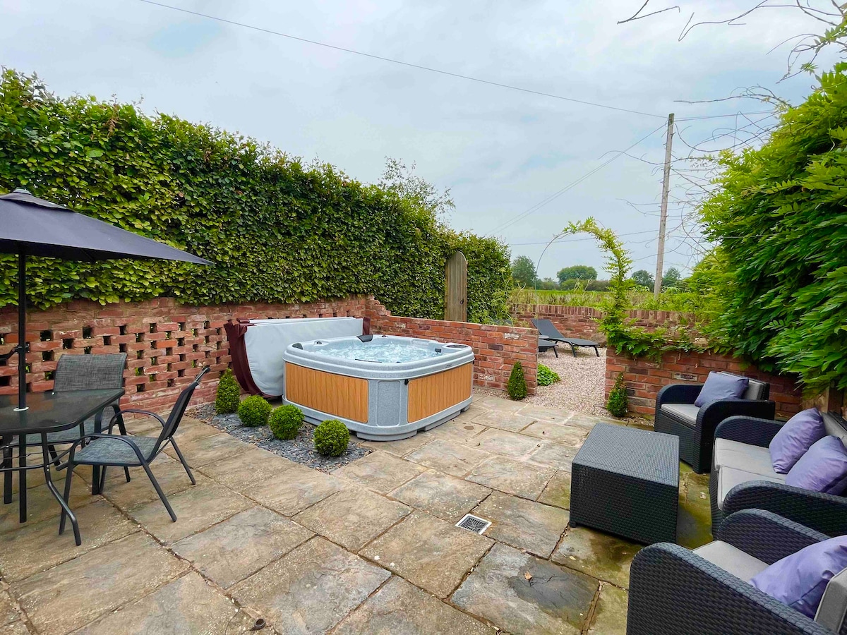 The private garden is shown, featuring a hot tub surrounded by greenery. Seating areas, including a lounge set and dining table with chairs, provide options for relaxation. Mature hedges offer seclusion, while paved stones create a neat and functional outdoor space.