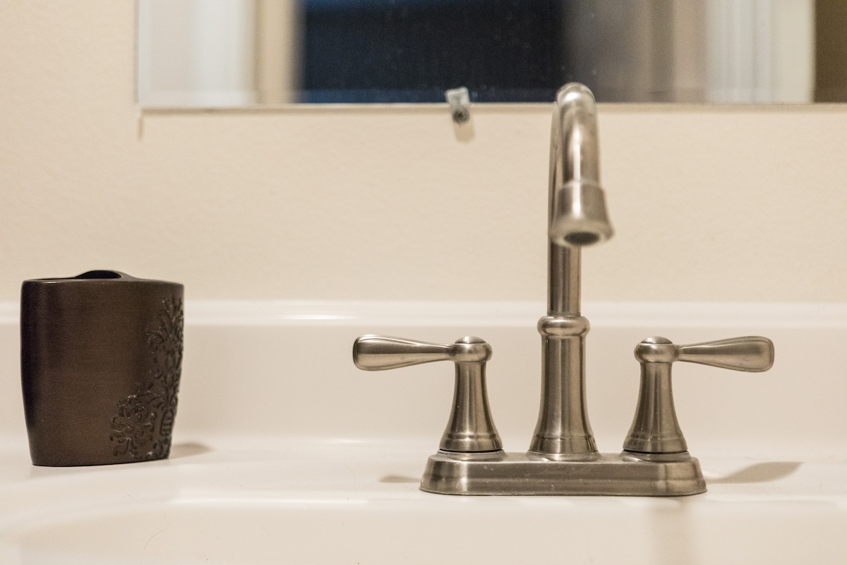 A sleek bathroom sink features a modern faucet with dual handles for temperature control. Next to the sink, a dark-toned decorative container adds a subtle touch of elegance. The space is well-lit, with a clear mirror reflecting the surroundings.
