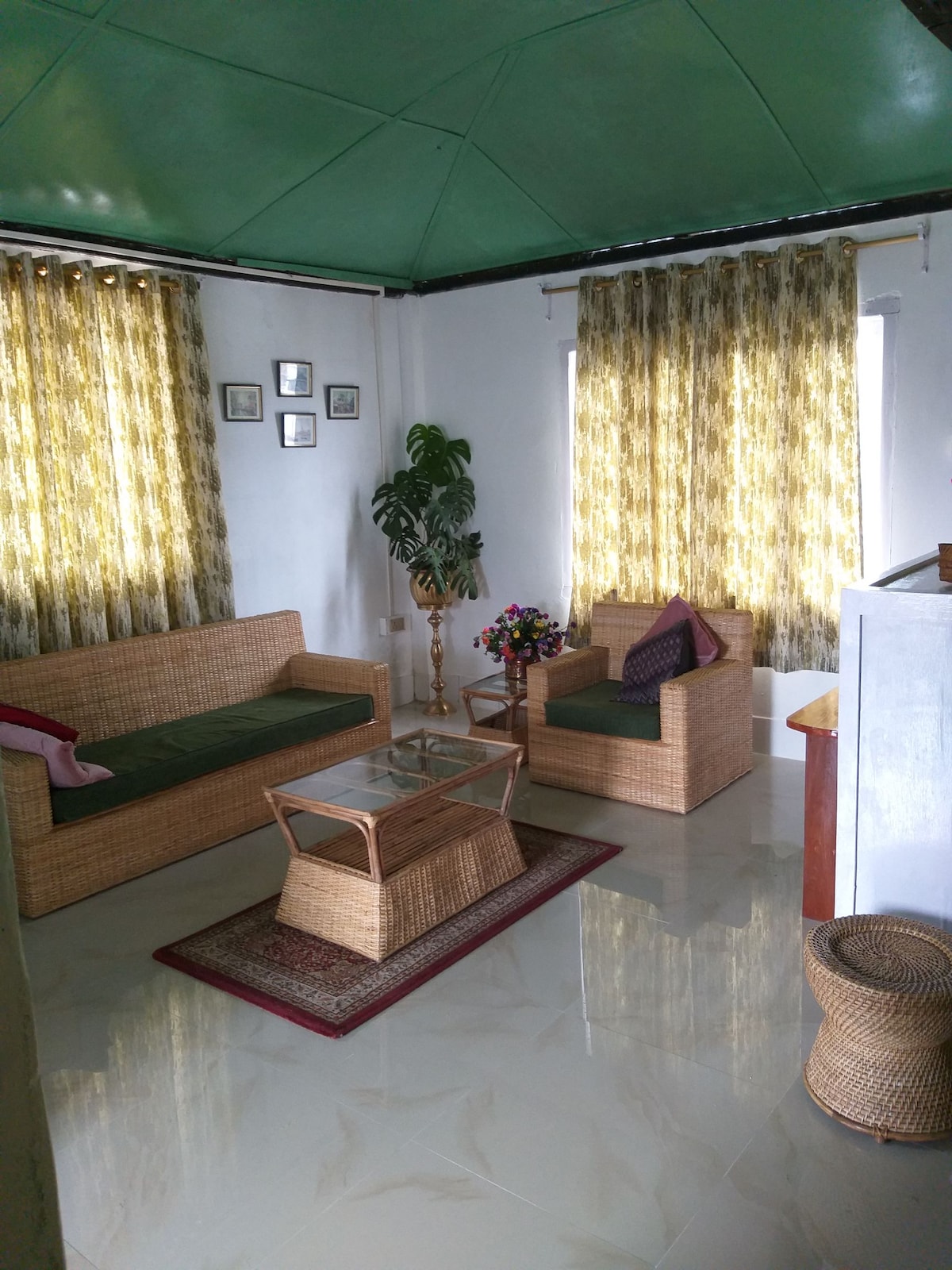 A bright living area features two woven seating arrangements, enhancing the room's spacious feel. A glass coffee table sits atop a decorative rug, complemented by a small flower arrangement. Natural light filters through patterned curtains, illuminating the warm colors of the furnishings.