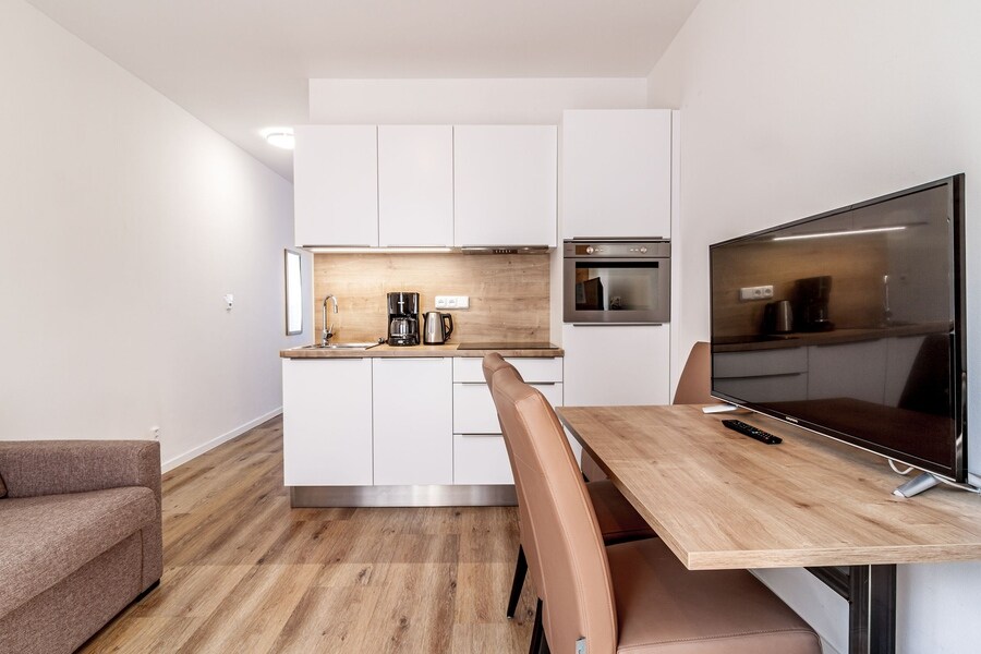 A modern kitchen area features sleek white cabinetry and wood accents. A coffee maker and kettle are visible on the countertop. A dining table with brown chairs is positioned next to a television, enhancing the open and functional layout.