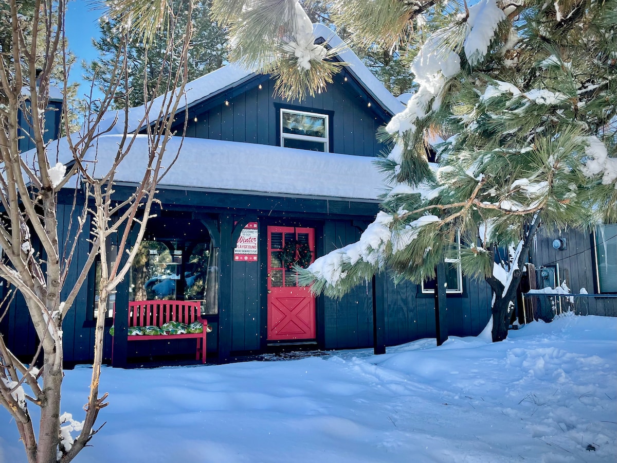 The exterior of a cabin is framed by snow-covered trees, showcasing a dark facade with bright red doors and trim. A welcoming red bench is positioned on the front porch, while a layer of fresh snow covers the ground.