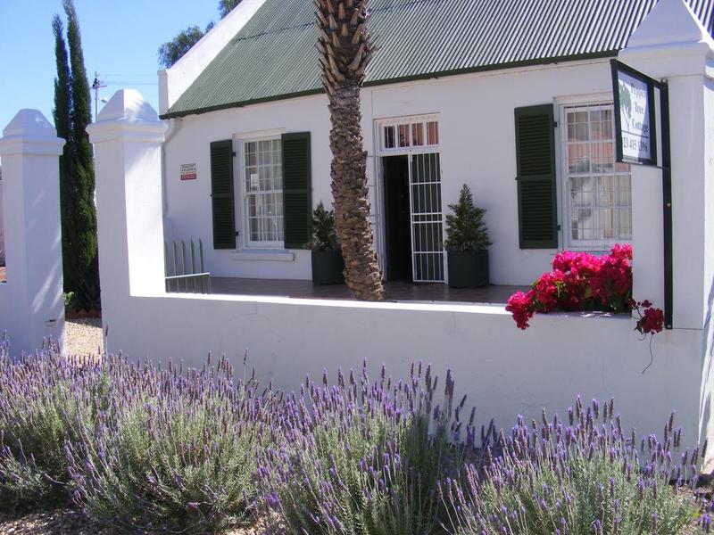 A charming white cottage is framed by lush lavender bushes and vibrant pink flowers. The green roof and shutters complement the exterior, while a palm tree adds a touch of nature. A gated entrance enhances security and accessibility.