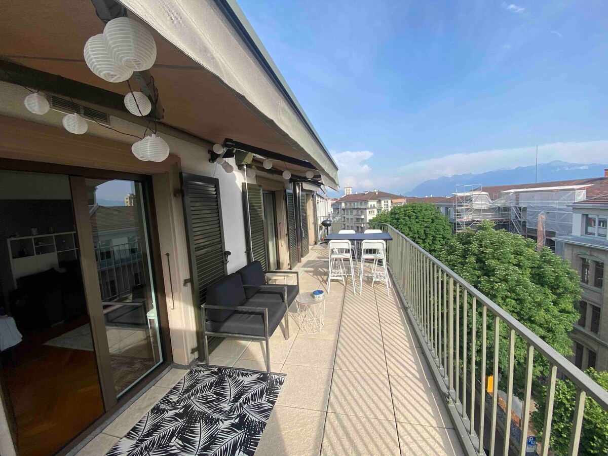 A spacious terrace is visible, featuring a railing and outdoor seating arrangements. Two high chairs and a small table invite relaxation, while views of trees and distant mountains create a serene backdrop. Neutral tones are complemented by decorative lanterns suspended above.