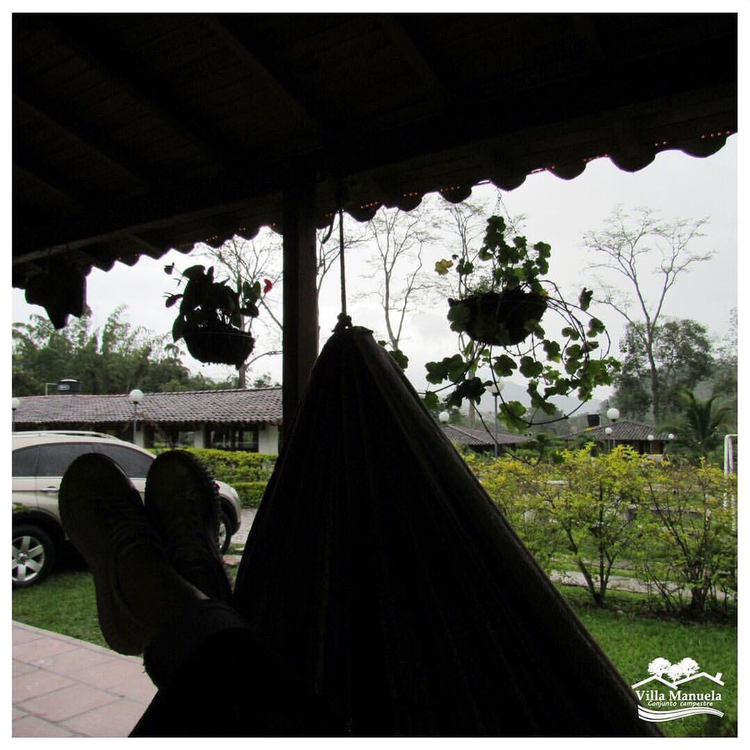 A cozy hammock is positioned under a covered porch, inviting relaxation. Planters with hanging greenery add a touch of nature, while a view of the surrounding landscape, including trees and the parking area, offers a serene atmosphere.