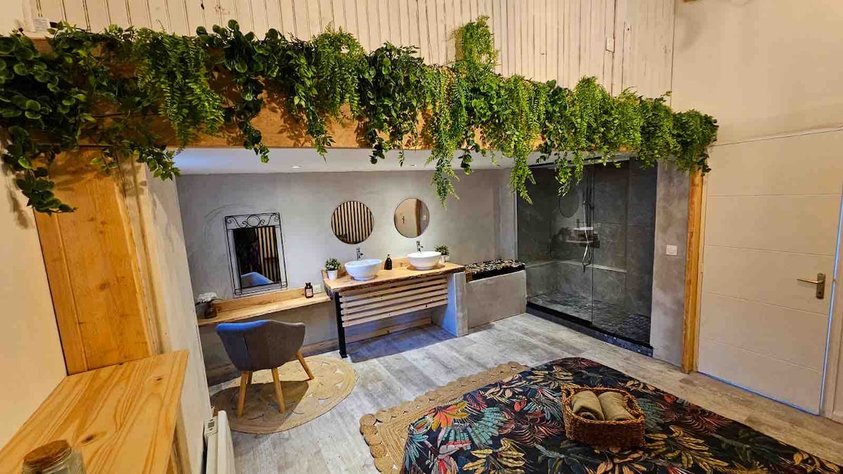 A modern suite features a spacious layout with natural wood accents and greenery overhead. Two round mirrors enhance a double vanity with ample storage. A large walk-in shower is visible alongside a decorative rug and a cozy bed adorned with foliage-inspired patterns.