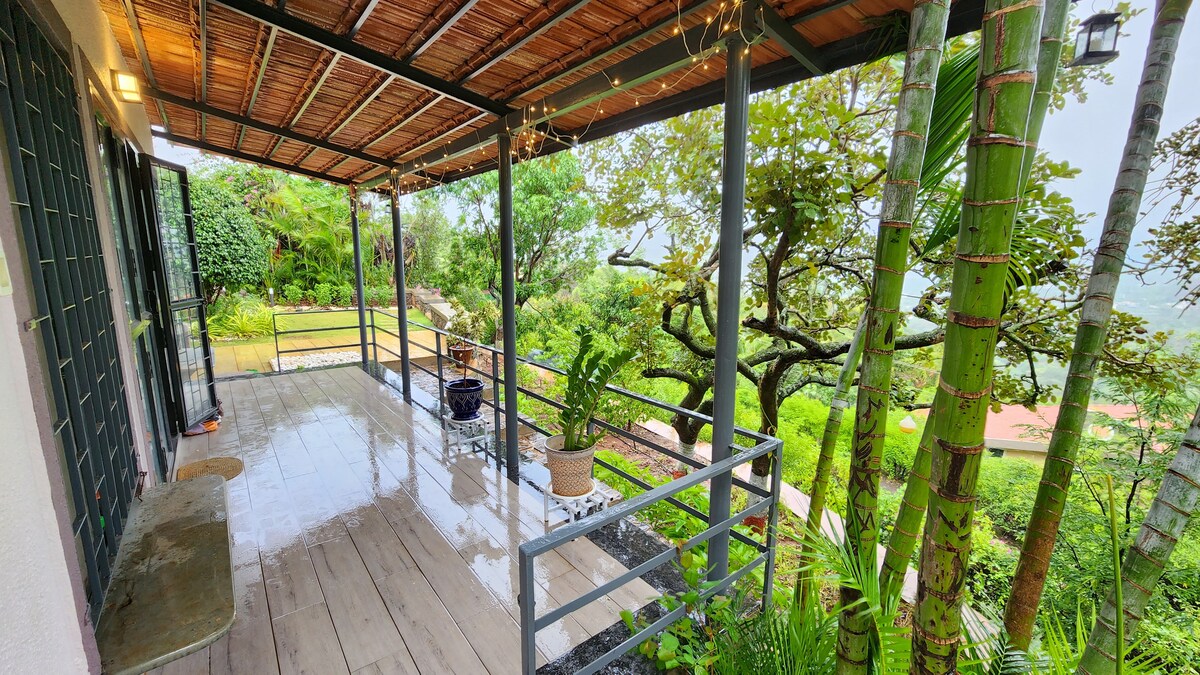 An outdoor patio area is presented, featuring a wooden floor and a railing along one side. Lush greenery, including palm trees and various plants, is visible in the surroundings. Subtle lighting adorns the structural beams, contributing to a serene and inviting ambiance.