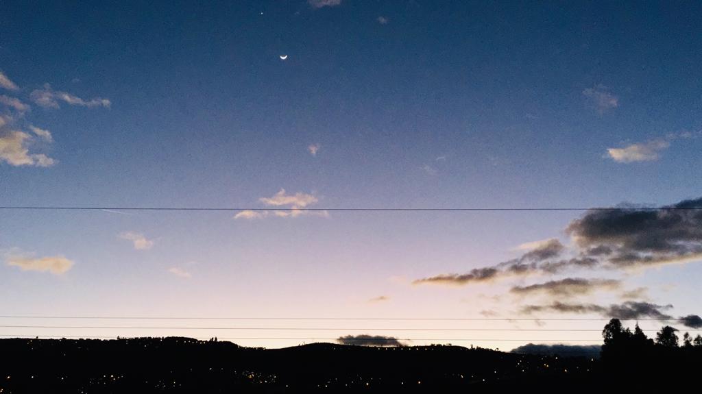 A serene nighttime sky transitions from deep blue to soft lavender, accented by wispy clouds. The crescent moon is visible, casting a gentle light over the distant landscape, including silhouettes of hills and glimmering city lights below.