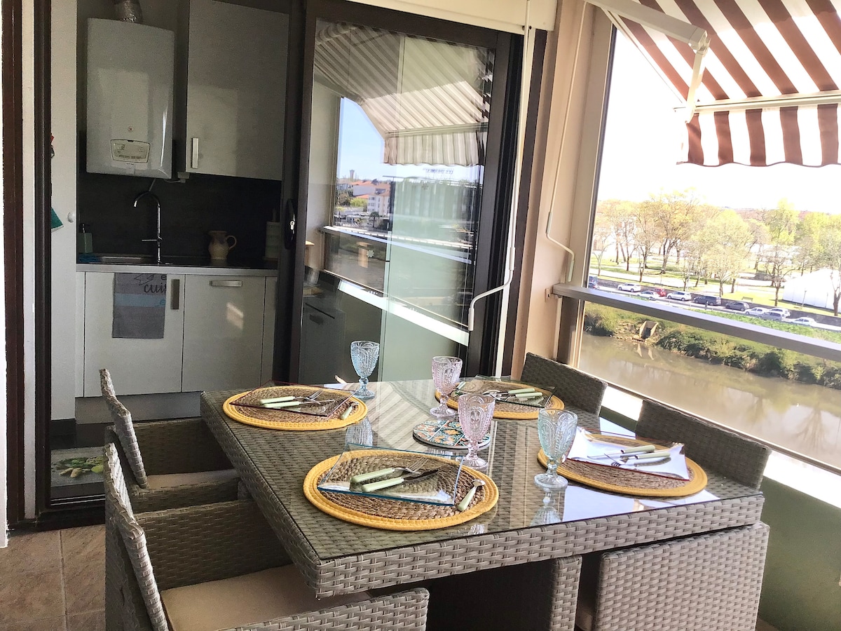 A dining area features a glass table surrounded by six wicker chairs, set beside large windows with striped shades. The adjoining kitchenette is visible, showcasing essential amenities. A scenic view of the Adour River is present outside, enhancing the inviting space.
