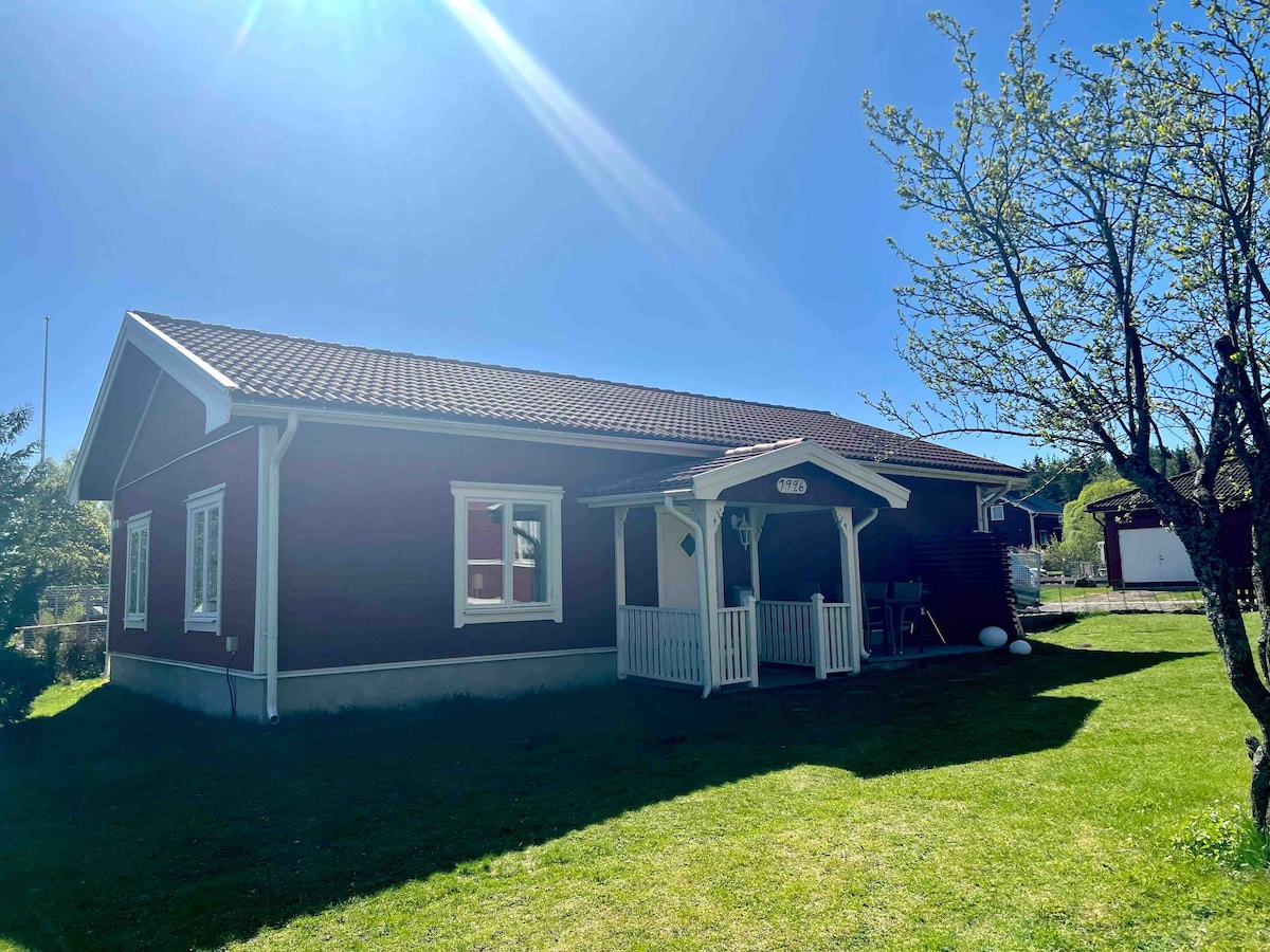 A red wooden house with a welcoming front porch is surrounded by a well-maintained lawn. Bright sunlight streams down, highlighting the structure's white window frames and roofing tiles. Nearby, a tree provides shade, enhancing the inviting outdoor space.