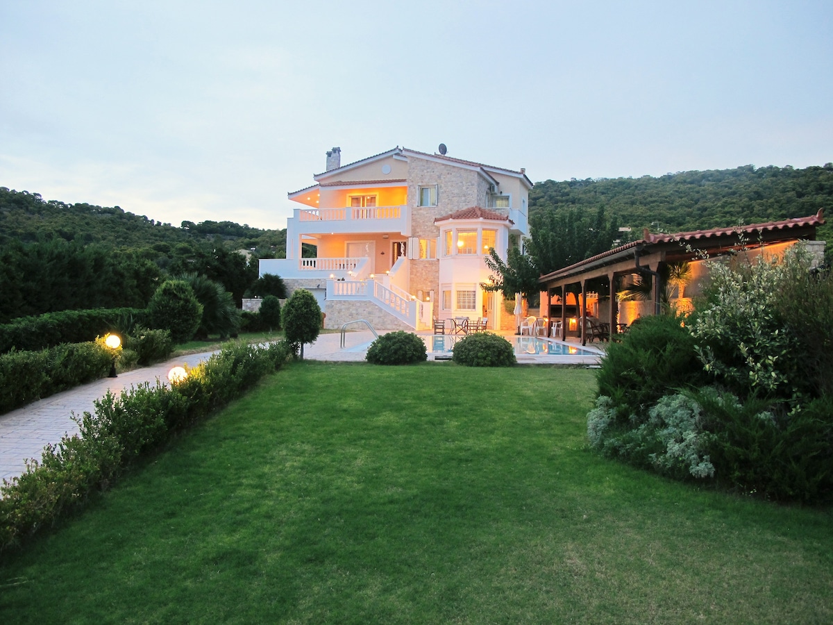 A spacious villa is surrounded by manicured lawns and greenery. The exterior features a combination of stone and light-colored walls, with expansive balconies. A covered outdoor area includes seating beside the swimming pool, inviting relaxation and social gatherings.