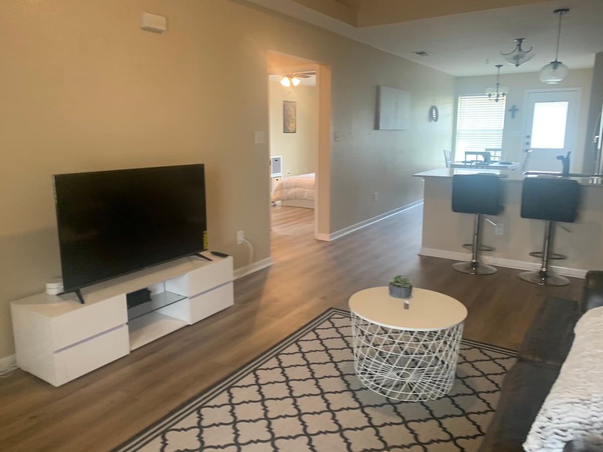 A cozy living area features a flat-screen TV positioned on a white media console. A round coffee table sits on a patterned rug, with two bar stools at a nearby countertop. Soft lighting and warm wood-like flooring enhance the inviting space.