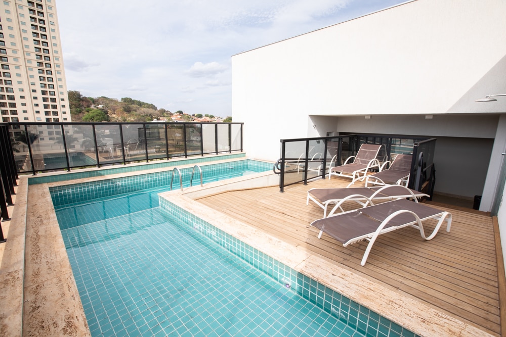 A rooftop swimming pool is bordered by a wooden deck, featuring several lounge chairs positioned for relaxation. The clear water reflects the sky, with views of the surrounding landscape visible in the background.
