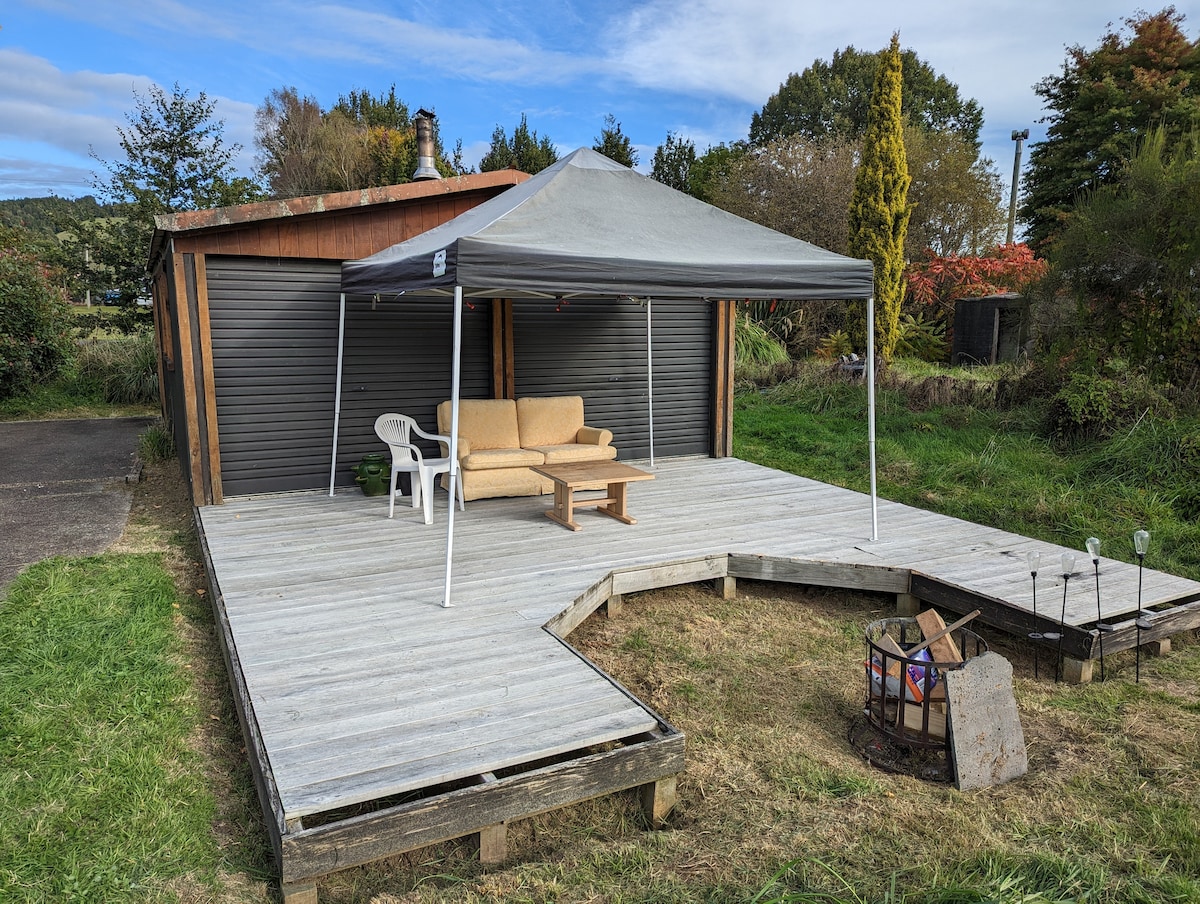 A sheltered seating area is shown on a wooden deck, featuring a comfortable couch and a small table. A fire pit is located nearby on the grass, surrounded by natural vegetation. A gray canopy provides shade, enhancing the outdoor setting.