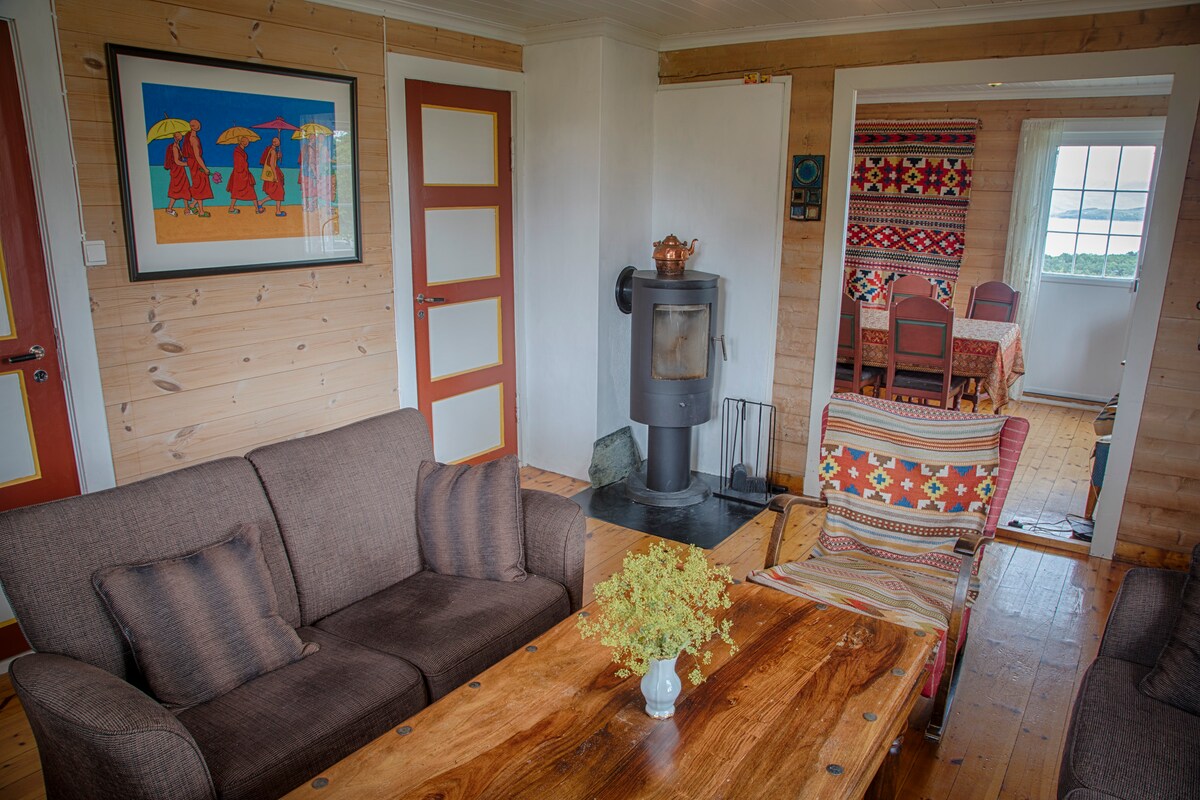 A cozy living area is presented with a wooden table at the center, surrounded by a pair of comfortable sofas. The wall features a framed artwork with three figures under umbrellas. A wood stove adds warmth, while a glimpse of the dining area is visible through an open doorway.