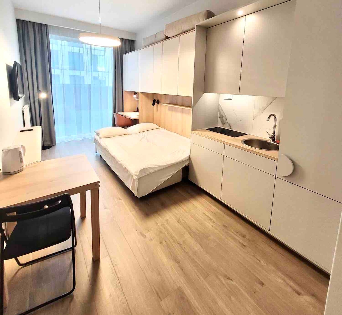 A compact studio space is showcased with a spacious bed and integrated kitchen area. A work desk and chair are present, along with a television mounted on the wall. Large floor-to-ceiling windows allow natural light to fill the room, enhancing the clean design.