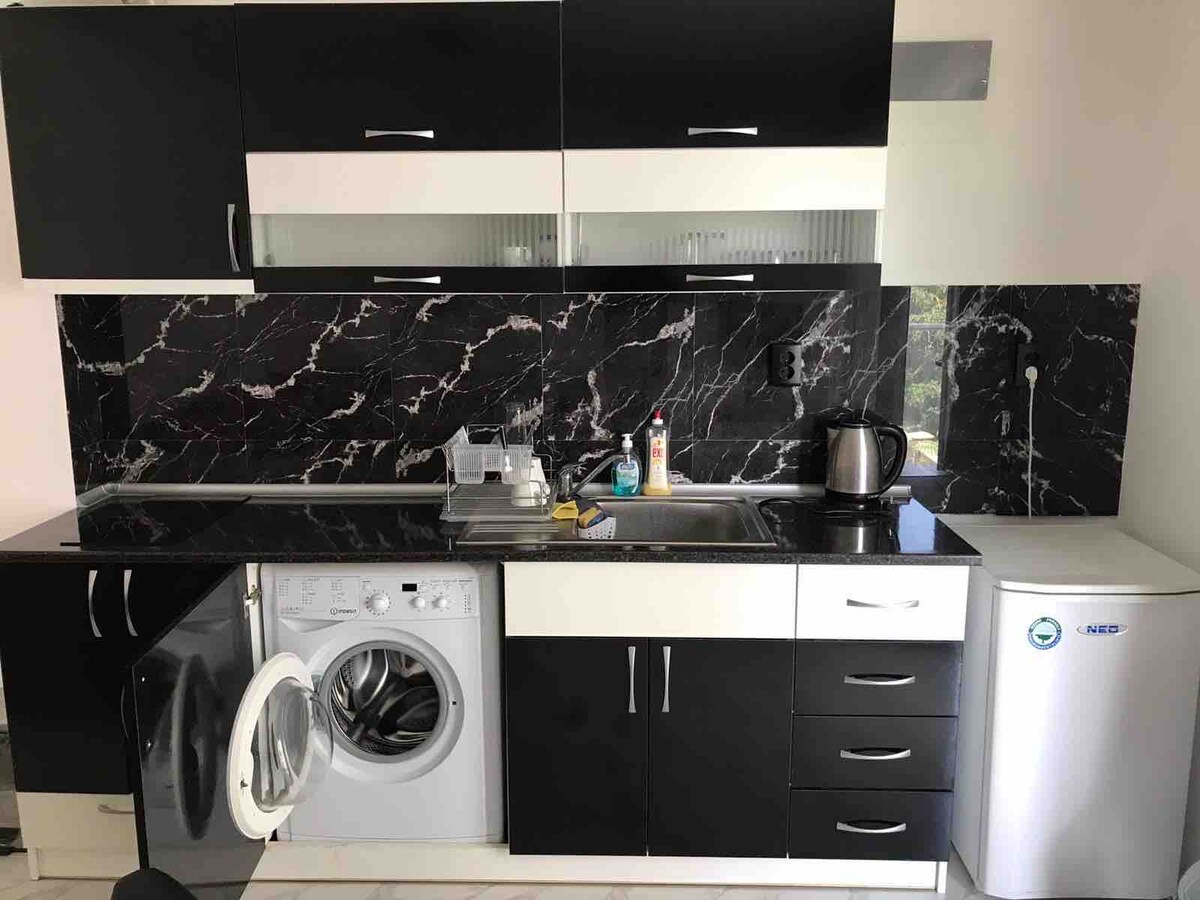 A modern kitchen is presented with sleek black cabinetry and a contrasting marble backsplash. A stainless steel kettle and a small white refrigerator are visible, alongside a washing machine with an open door. A sink and a variety of kitchen utensils are neatly arranged.