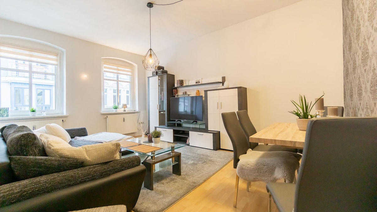 A bright and airy living area features a spacious sofa paired with a decorative coffee table. A wall-mounted flat-screen TV is placed on a contemporary unit. Natural light filters through large windows, and a dining table with chairs completes the inviting space.