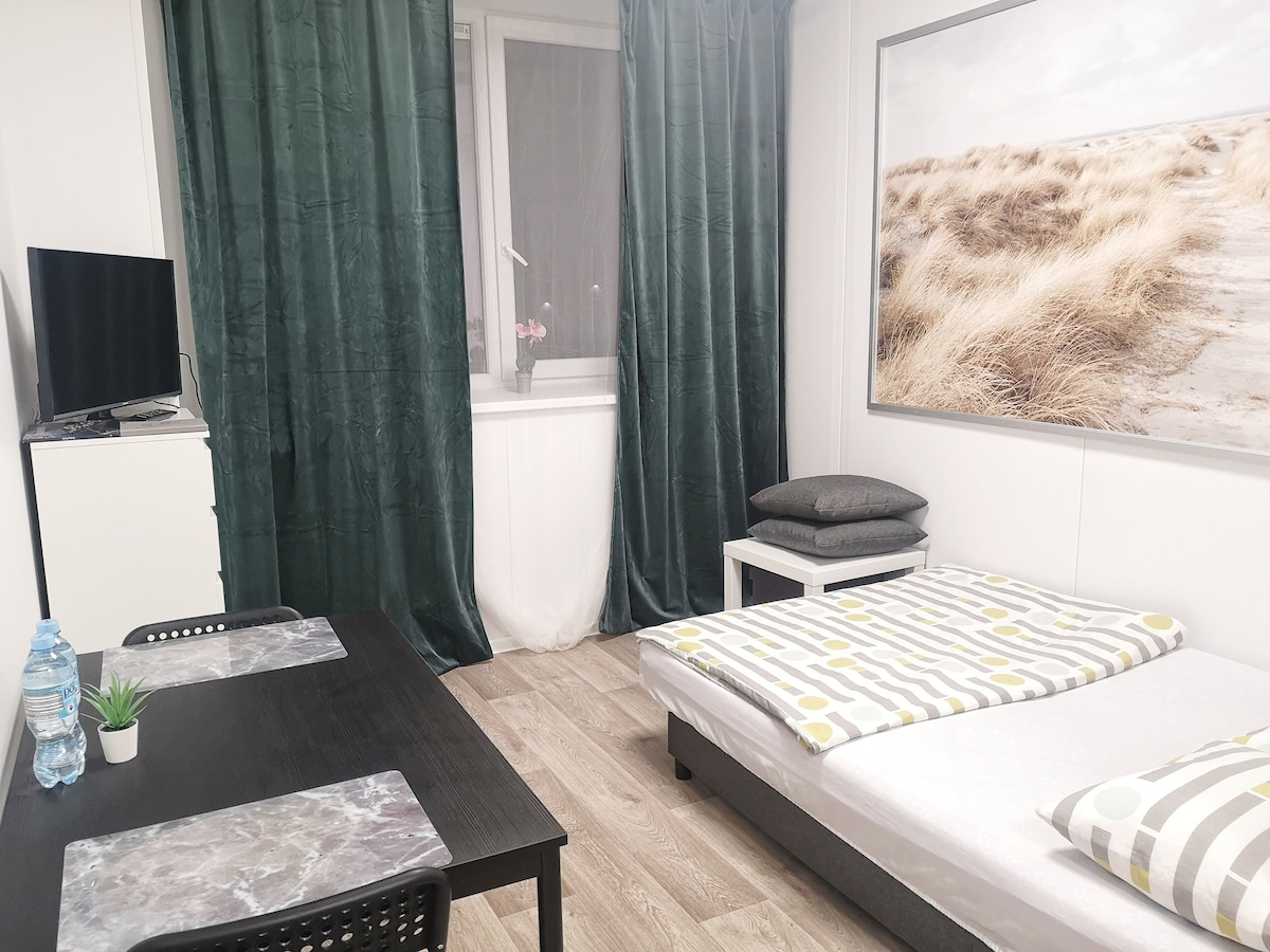 A studio apartment is shown with a spacious bed dressed in neutral linens. A dining area with two chairs is visible, accompanied by a small plant on the table. A large window is framed by green curtains, enhancing the light and setting.