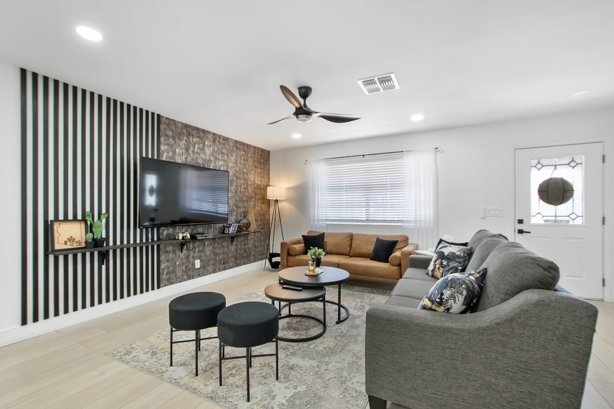 A modern living room is presented, featuring a comfortable sectional sofa and a distinctive brown leather couch. A sleek television is mounted on a textured wall. Circular coffee tables are positioned on a light rug, and natural light filters through sheer curtains at the window.