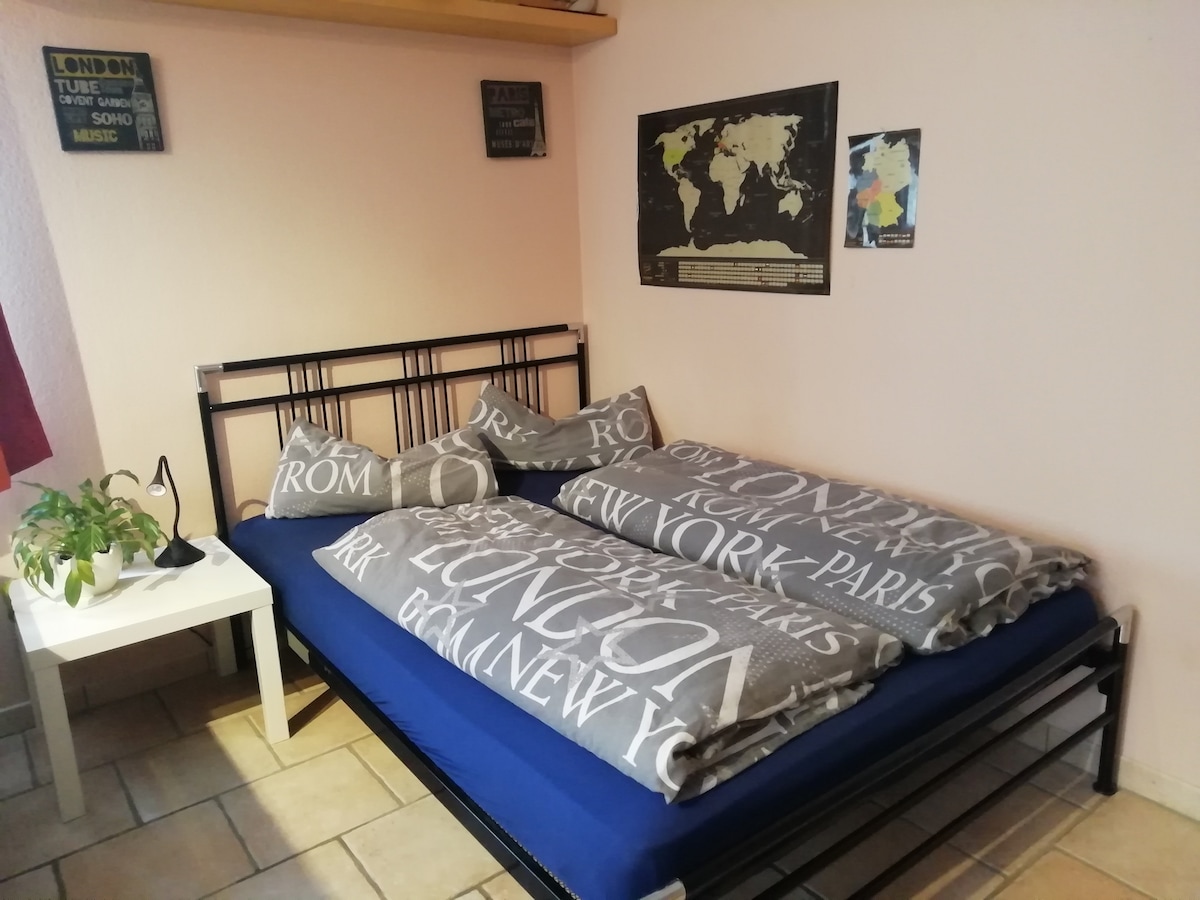 A spacious bed with cozy blankets and decorative pillows is positioned against a light pink wall. A bedside table features a small plant, while wall art, including a world map and signs, adds visual interest to the room.
