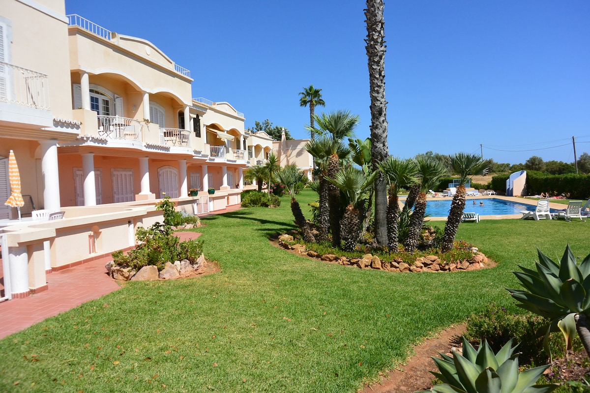 A well-maintained garden surrounds the private swimming pool, featuring lush greenery and palm trees. The building's balconies provide access to the ground floor apartments, while sun loungers are arranged around the pool area, creating a calm and inviting atmosphere.