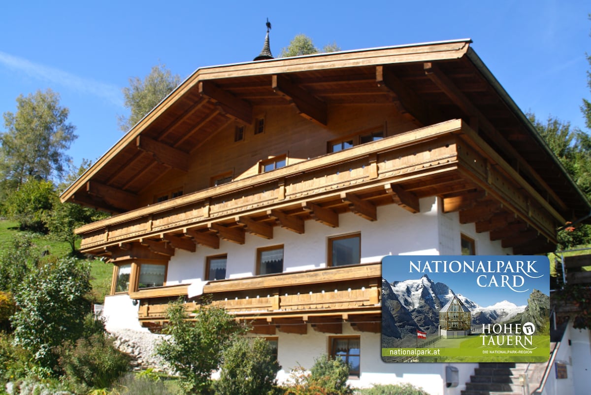 A traditional alpine-style chalet with a wooden exterior and large balconies showcases stunning craftsmanship. The structure features multiple levels, surrounded by greenery and trees, highlighting a serene mountain setting. The Nationalpark SommerCard logo is visible in the foreground.