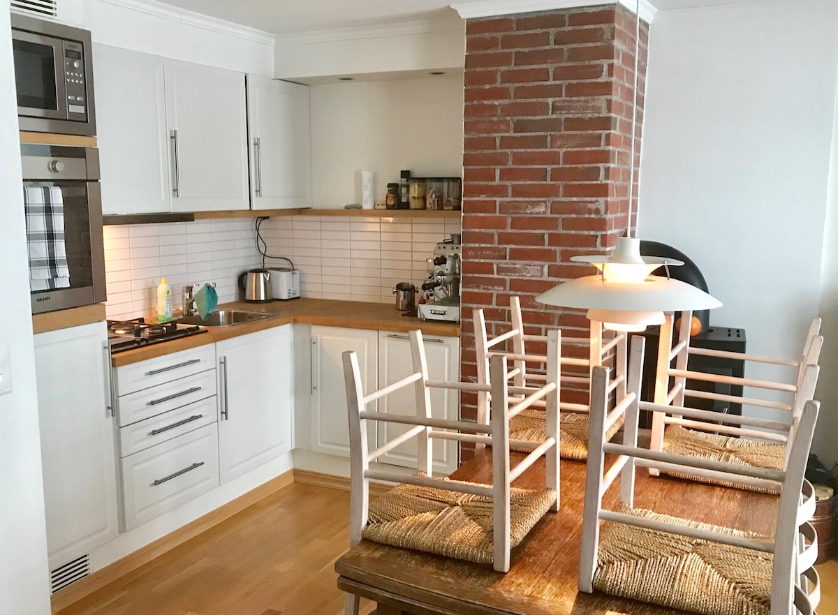 A modern kitchen space is shown, featuring white cabinetry and wooden countertops. Kitchen appliances are neatly arranged, including a stovetop and a kettle. A rustic wooden dining table with woven chairs stacked upside down is visualized, with a standing lamp gently illuminating the area.