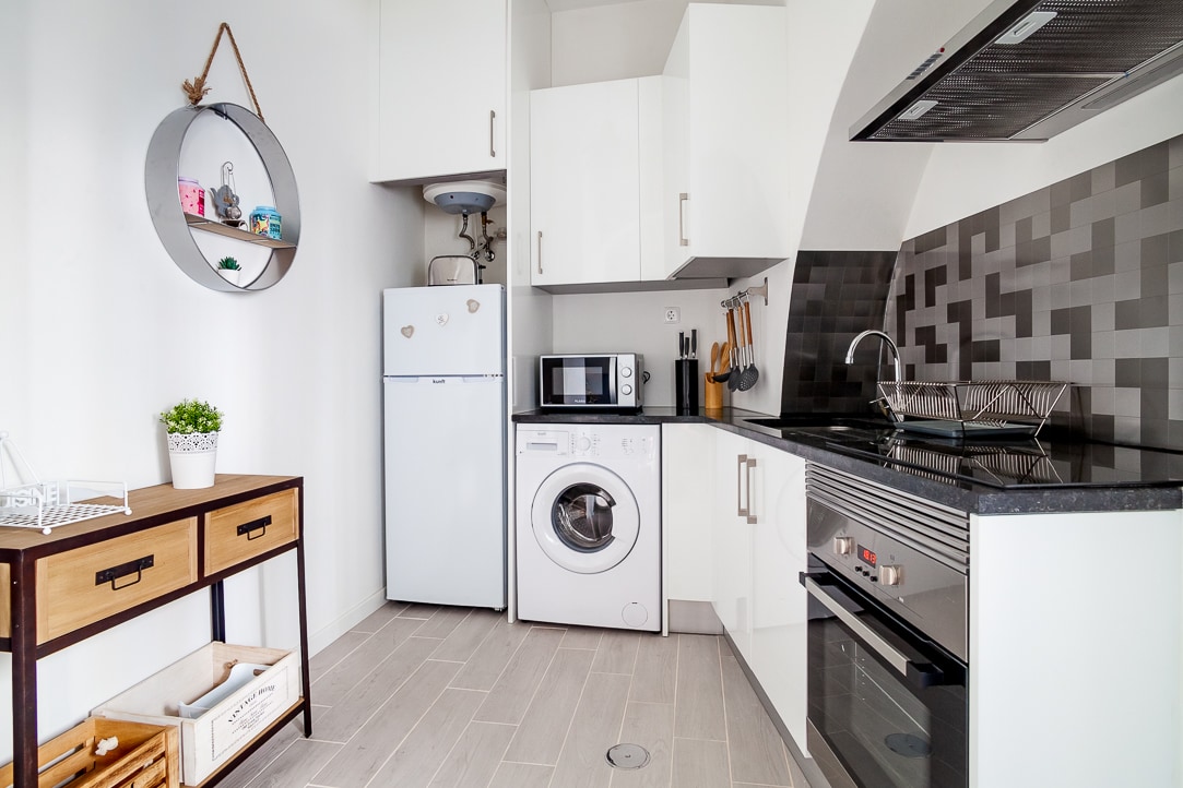 The kitchen is equipped with modern appliances, including a refrigerator, microwave, and oven. A washing machine is also present. Light-colored cabinetry complements the contrasting dark countertops. Decorative elements are displayed on a round shelf, while a wooden console adds functional storage.
