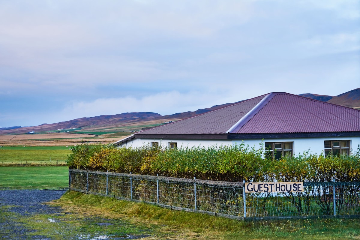 A guest house with a red gabled roof is set against a backdrop of rolling hills. The building is surrounded by a low fence and vibrant greenery. A sign stating 'GUEST HOUSE' is prominently displayed in front.