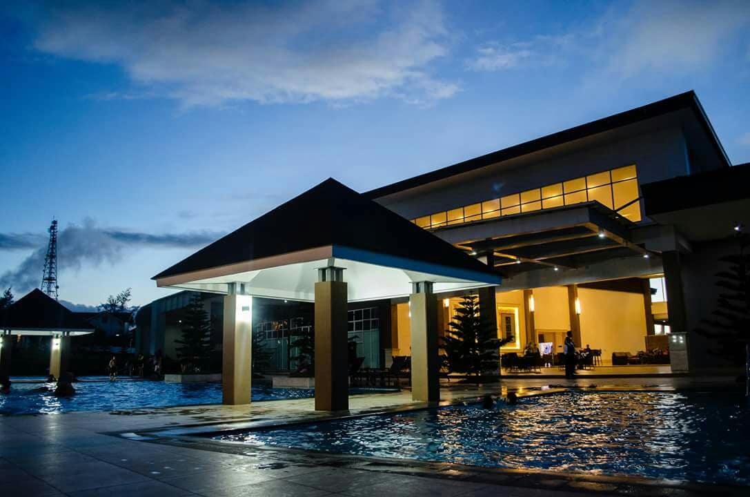 A modern clubhouse features a spacious outdoor pool area, with illuminated structures complementing the night sky. The water’s surface reflects the soft glow of lights, creating a serene atmosphere. Nearby seating areas are visible, providing a relaxing space for guests.