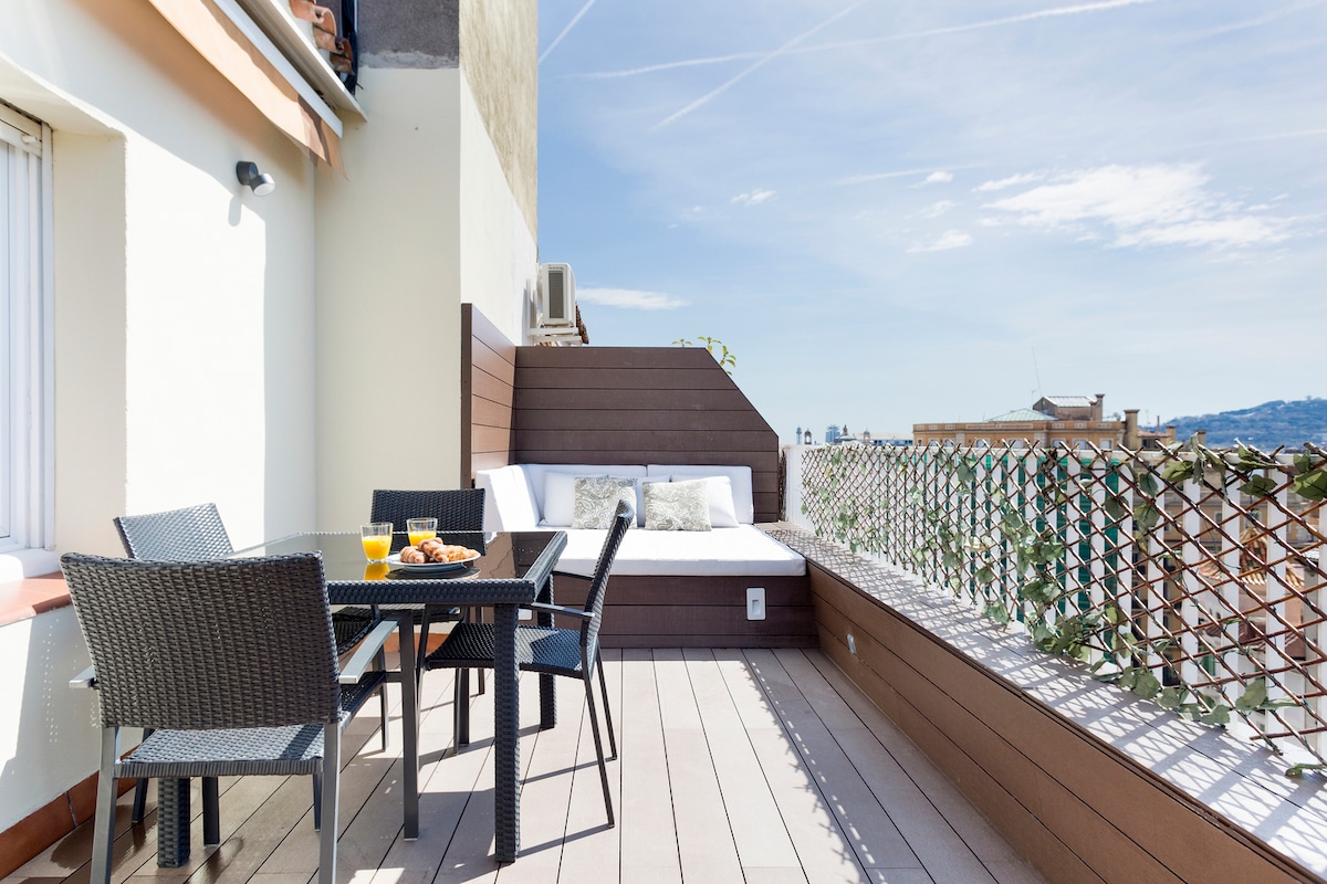 An expansive balcony is presented, featuring a table with four chairs and a cozy lounge area. A set of breakfast items is seen on the table, while the lounge space is outfitted with cushions against a wooden backdrop. The surrounding fence includes climbing plants and offers a city view.