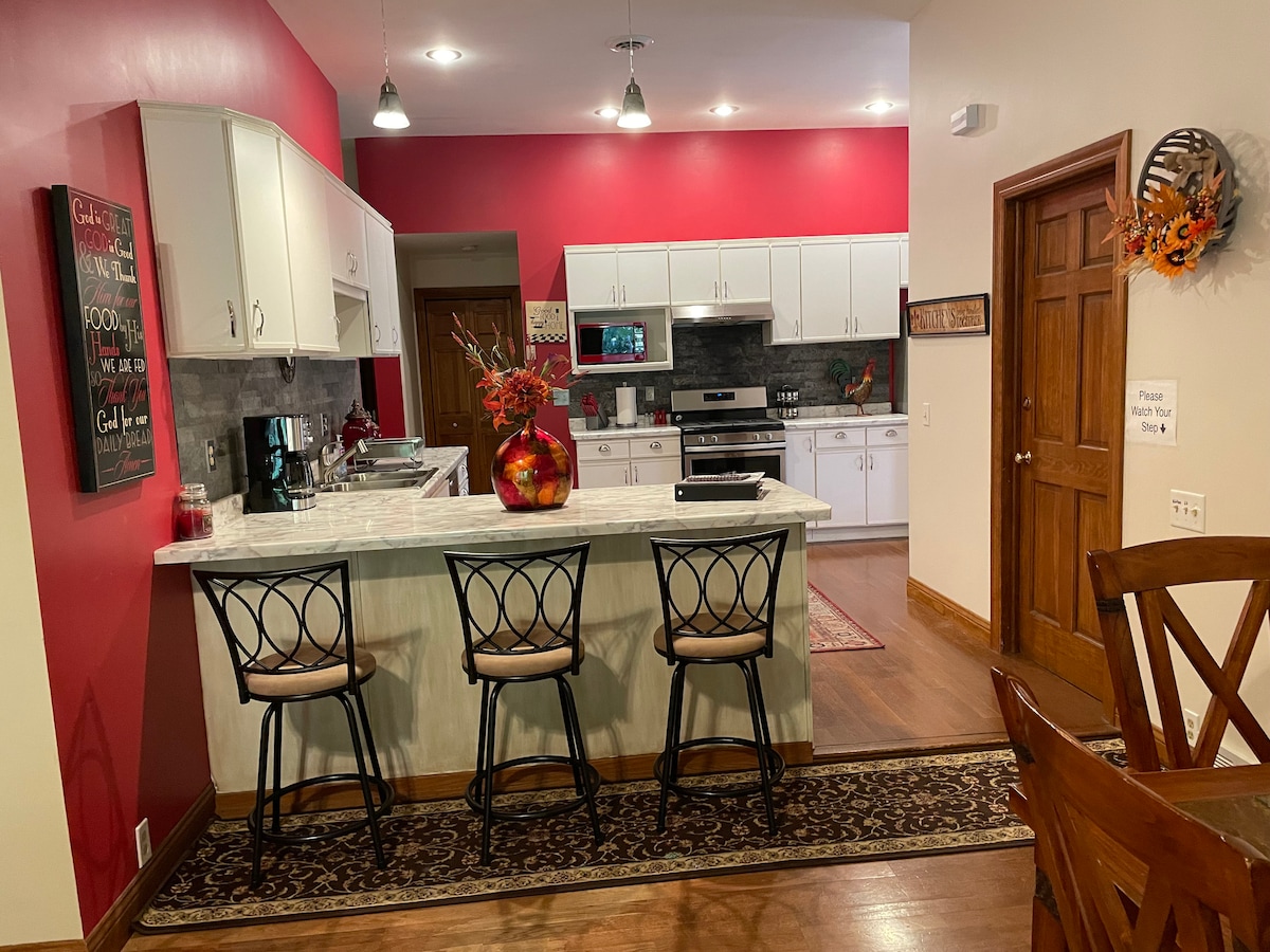 A modern kitchen features red accent walls and a large central island with seating for three. Stainless steel appliances are situated against a contemporary backsplash, while natural light brightens the space. A decorative vase with flowers sits prominently on the island.