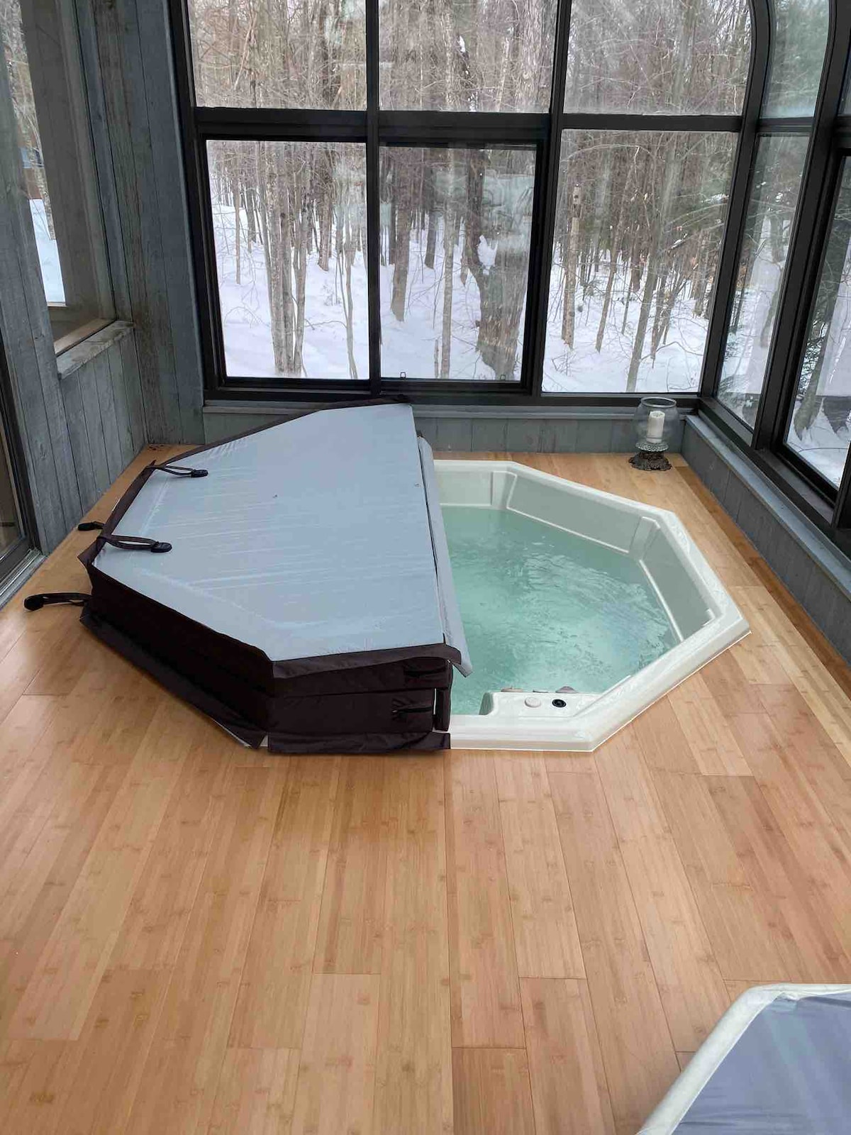 A spa area features a light-colored, octagonal hot tub with a protective cover resting on the edge. Large windows surround the space, offering views of the snow-covered landscape outside. The bamboo flooring enhances the serene ambiance.
