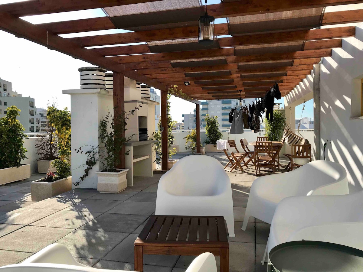 An outdoor terrace is featured, furnished with modern white lounge chairs and wooden tables. A barbecue area is visible, surrounded by greenery. Clothes are hung on a line, and sunlight casts shadows across the tiled surface, creating a relaxed ambiance.