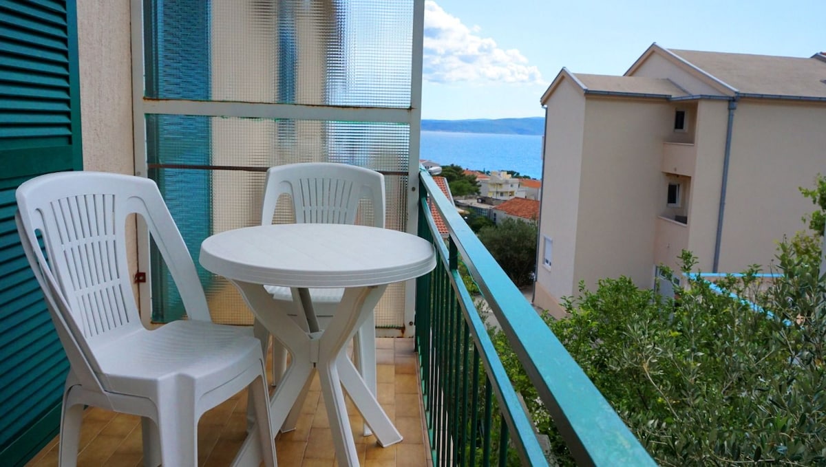 A small balcony features a round table accompanied by two white chairs. A view of the sea is visible in the background, complemented by neighboring buildings and greenery in the foreground.