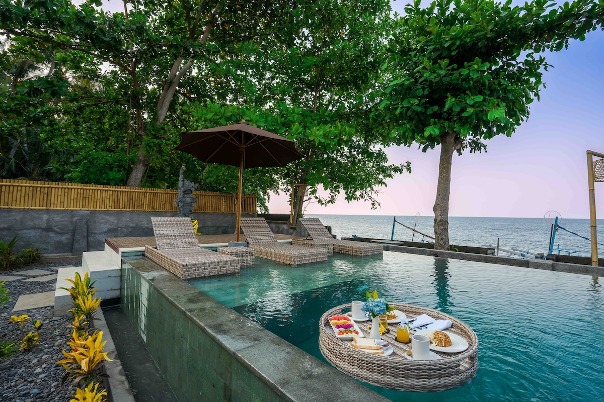 A serene pool area is surrounded by lush greenery, with loungers positioned under an umbrella for shade. Floating trays display a breakfast spread while the tranquil ocean is visible in the background, offering an inviting space for relaxation.