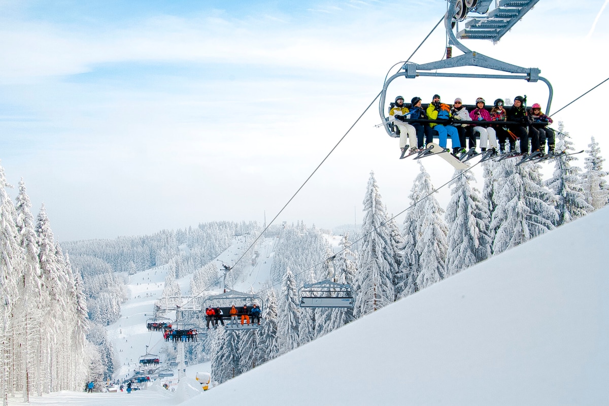 A chairlift carries multiple skiers over a snow-covered landscape, with dense evergreen trees lining the hills. The snowy slopes extend into the distance, showing a winter wonderland of white against a bright sky.