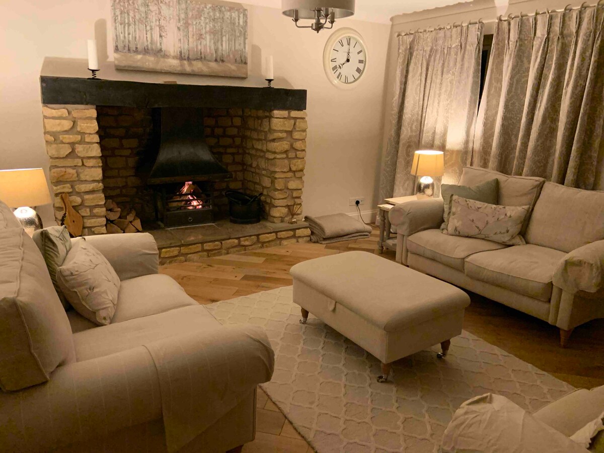 The cozy living area showcases a stone fireplace with a gently flickering fire. Light-colored sofas surround a central ottoman on a patterned rug. Soft curtains frame the windows, while warm lamps provide a soothing glow. A clock and artwork add subtle decorative elements.