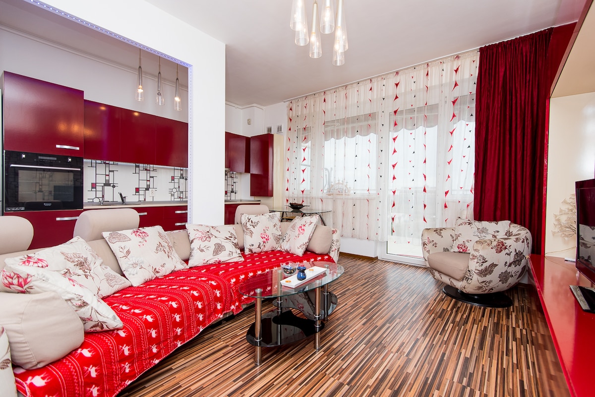 A modern living area with a plush, curvy sofa upholstered in floral fabric, complemented by a vibrant red throw. A glass coffee table is positioned at the center, and large windows draped in light fabric allow natural light to fill the space.